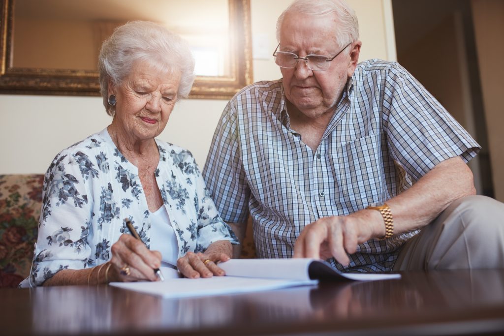 Indoor shot of mature couple at home signing documents together. Senior man and woman sitting on sofa doing retirement paperwork.